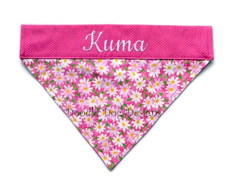 May include: Pink dog bandana with a white daisy print and a pink polka dot border. The bandana is personalized with the name "Kuma" in white embroidery.