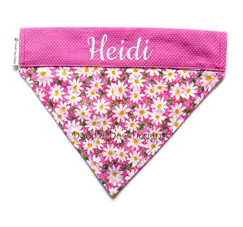 May include: Pink dog bandana with white daisy print and white polka dots. The bandana has the name "Heidi" embroidered on the top.