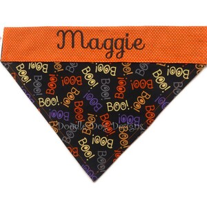 Puede incluir: Un pa&ntilde;uelo para perro con tem&aacute;tica de Halloween con el nombre "Maggie" bordado en una banda naranja con lunares. La secci&oacute;n principal es negra con "BOO!" en varios colores. El pa&ntilde;uelo es triangular.