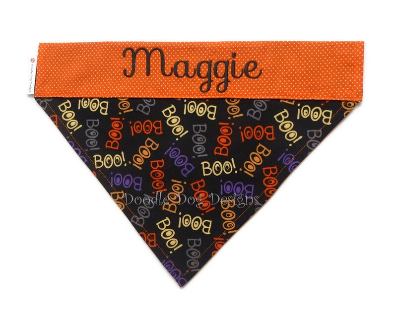 Puede incluir: Un pa&ntilde;uelo para perro con tem&aacute;tica de Halloween con el nombre "Maggie" bordado en una banda naranja con lunares. La secci&oacute;n principal es negra con "BOO!" en varios colores. El pa&ntilde;uelo es triangular.