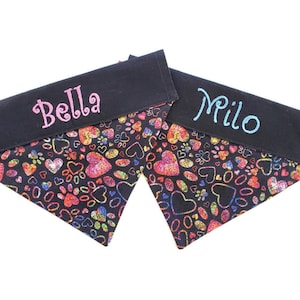 May include: Two black dog bandanas with a colorful heart and paw print pattern. One bandana is embroidered with the name "Bella" in pink and the other with the name "Milo" in blue.