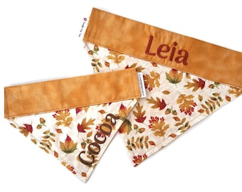 Puede incluir: Dos pañuelos para perros con un estampado de hojas de otoño. Los pañuelos tienen una parte superior marrón y un fondo blanco con hojas rojas, naranjas y verdes. Un pañuelo dice "Cocoa" y el otro dice "Leia".