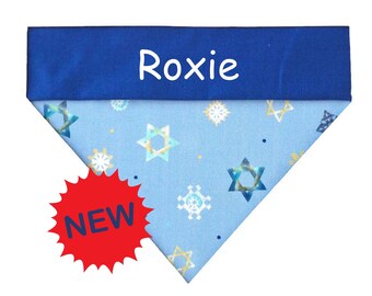 Puede incluir: Un pañuelo triangular para mascotas con una banda superior azul oscuro que lleva el texto blanco Roxie. La sección inferior azul claro muestra un patrón repetido de copos de nieve blancos y estrellas de seis puntas azules y doradas, con una etiqueta roja que dice NEW.
