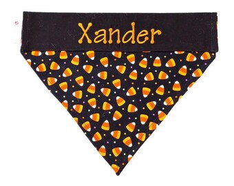 May include: A black and orange Halloween-themed dog bandana with a candy corn pattern. The bandana is triangular, with the name "Xander" embroidered in orange thread across the top. The candy corn design features orange, yellow, and white candies.