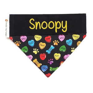 May include: Black dog bandana with a colorful pattern of hearts and paw prints. The hearts have various dog-related words on them, such as "woof", "bark", "play", and "fetch". The bandana has a black band at the top with the name "Snoopy" embroidered in yellow.
