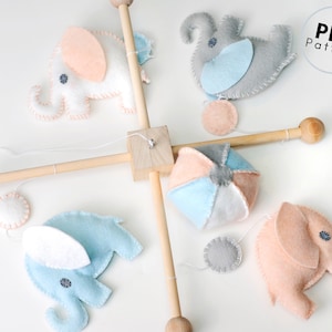 Può includere: Un mobile in legno con quattro giocattoli di elefante in feltro e una palla in feltro. Gli elefanti sono in tonalità di rosa, blu e grigio. La palla è multicolore con sfumature di blu, rosa e grigio. Il mobile è appeso a una base in legno.  Modello PDF.
