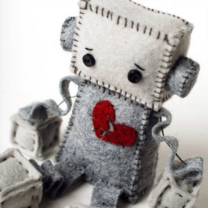 May include: A gray felt robot with a broken heart. The robot has a stitched-on face with black eyes and a stitched-on mouth. The robot is holding two gray felt blocks in its hands.