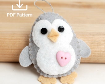 May include: A gray and white felt penguin ornament with black eyes, an orange beak, and a pink heart-shaped button on its chest.  The ornament has a loop for hanging.  PDF Pattern.