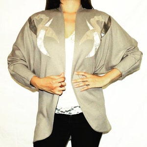 May include: A beige cardigan with a long, open front and long sleeves. The cardigan has a unique design with two silver and beige shapes on the front.