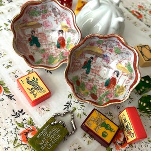May include: Two scalloped porcelain bowls with hand-painted scenes of figures in a garden, featuring gold-trimmed edges. Scattered around are various Mah Jongg tiles, including a green keychain reading MEMBER OF NATIONAL MAH JONGG LEAGUE.