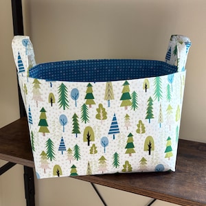 May include: A white fabric storage basket with blue handles and a green and blue tree pattern. The basket is sitting on a wooden shelf.