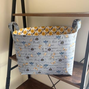 May include: A fabric storage basket with handles. The exterior is light blue with a floral and butterfly pattern and inspirational text. The interior lining features a yellow floral design. The basket sits on a wooden shelf.