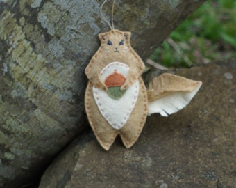 May include: A felt squirrel ornament with a brown body, white belly, and a brown acorn in its paws. The ornament has a loop for hanging.