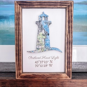 May include: A framed artwork featuring a lighthouse silhouette filled with a map design. The lighthouse is blue and white, set against a white background, and framed in dark wood. Text reads "Portland Head Light" and coordinates.