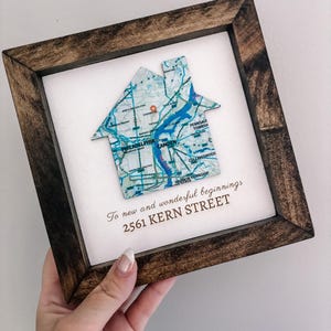 May include: A framed artwork featuring a house-shaped map cutout. The map depicts a city with blue waterways and roads. The frame is dark brown, and the artwork includes the text "To new and wonderful beginnings 2561 KERN STREET."