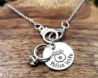 May include: Silver necklace with a police badge charm that says "Police Wife" and a ring charm. The necklace is on a wooden surface.