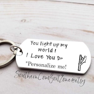 Op de afbeelding: Zilveren sleutelhanger met een boodschap die luidt "You light up my world! I Love You" met een hartje en een silhouet van een persoon die een elektriciteitspaal beklimt. De tekst "*Personalize me!" is ook inbegrepen.