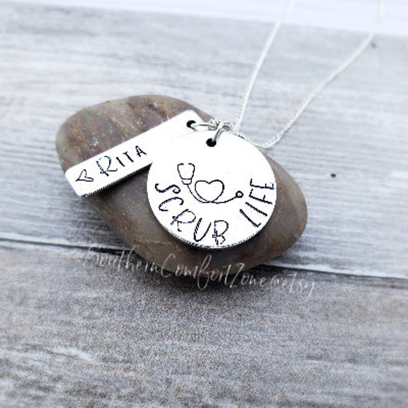 Op de afbeelding: Zilveren ketting met twee bedels. De grotere bedel is rond en zegt "Scrub Life" met een stethoscoop en een hart. De kleinere bedel is een rechthoekig label dat "BRITA" zegt.
