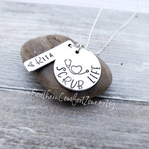 Op de afbeelding: Zilveren ketting met een ronde hanger waarop "Scrub Life" staat met een stethoscoop en een hart. Een kleinere tag met de naam "BRITA" hangt aan de ketting.