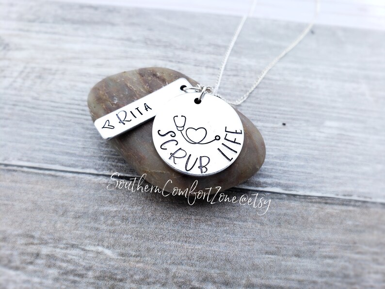 Op de afbeelding: Zilveren ketting met een ronde hanger waarop "Scrub Life" staat met een stethoscoop en een hart. Een kleinere tag met de naam "BRITA" hangt aan de ketting.