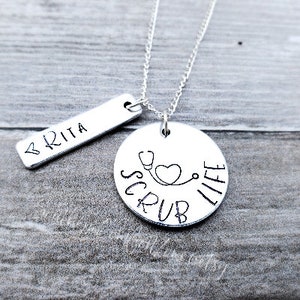 Op de afbeelding: Zilveren ketting met een ronde hanger waarop "Scrub Life" staat met een stethoscoop en een hart. Er is ook een kleinere staafhanger met de naam "BRITA" bij inbegrepen.