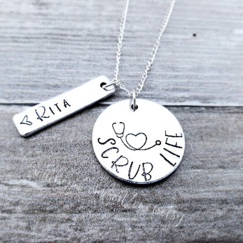 Op de afbeelding: Zilveren ketting met een ronde hanger waarop "Scrub Life" staat met een stethoscoop en een hart. Er is ook een kleinere staafhanger met de naam "BRITA" bij inbegrepen.