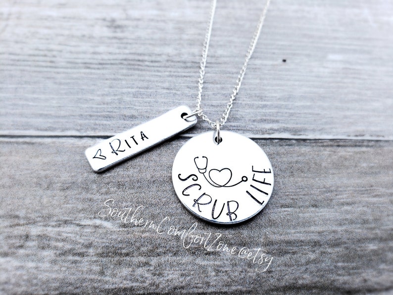 Op de afbeelding: Zilveren ketting met een ronde hanger waarop "Scrub Life" staat met een stethoscoop en een hartontwerp. Een kleinere staafhanger zegt "BRITA".