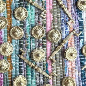 May include: An assortment of gold-colored cabinet hardware, including round knobs and rectangular pulls, arranged on a colorful, striped rug. The hardware has a vintage or antique aesthetic, with detailed designs.