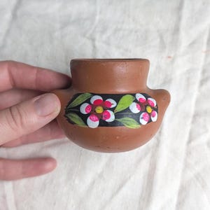 May include: A small, brown ceramic pot with a floral design. The pot features a band of flowers with red and white petals, green leaves, and a black background. The pot is held in a hand.