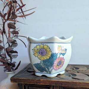 Op de afbeelding: Een kleine, witte keramische plantenbak met een geschulpte rand. De plantenbak is versierd met een bloemenontwerp met gele en roze zonnebloemen, groene bladeren en een blauwe rand. De plantenbak staat op een donkere houten kist.
