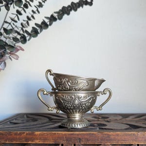 May include: Two antique silver serving dishes. The bottom dish is a handled bowl with a pedestal base. The top dish is a small gravy boat. Both dishes have ornate floral and scrollwork designs. The dishes are stacked on a dark wooden cabinet.