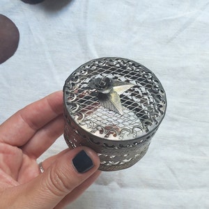 Puede incluir: Una caja de metal plateado para guardar peque&ntilde;os objetos con un dise&ntilde;o decorativo e intrincado. La tapa presenta un patr&oacute;n de malla y una peque&ntilde;a flor dorada con un centro en forma de estrella.