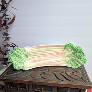 Puede incluir: Una fuente de cerámica con forma de tallo de ruibarbo. La fuente es principalmente blanca con detalles en rosa y verde, y presenta bordes detallados en forma de hojas. La fuente está colocada sobre un cofre de madera oscura.