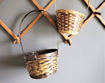 Puede incluir: Dos cestas tejidas con asas colgadas de un estante de pared de madera. Las cestas están hechas de materiales naturales y tienen una combinación de colores marrón claro y marrón oscuro.