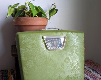 May include: A vintage Borg bathroom scale in a vibrant olive green color featuring an embossed damask pattern across the surface. The scale has a central analog display window with a silver frame, showing numbers and the brand name Borg.