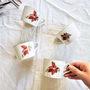 Puede incluir: Cuatro tazas de cerámica blanca con un diseño de flor de pascua. Cada taza presenta una flor de pascua roja y verde con un borde verde y un asa blanca. Las tazas están dispuestas sobre una tela blanca.
