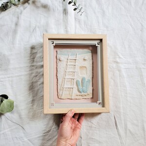 May include: A framed 3D artwork depicting a white adobe house with a ladder and a cactus in front. The artwork is set against a pink background and has a light wood frame.