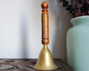 May include: A brass bell with a wooden handle. The bell is sitting on a wooden table with carved details.