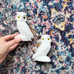 Op de afbeelding: Twee witte keramische uilbeeldjes met gele ogen op een kleurrijk, gepatroneerd tapijt.