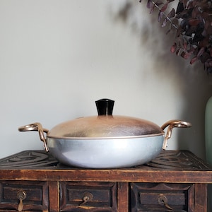May include: A vintage copper and aluminum pot with a black knob on the lid. The pot is sitting on a wooden table with drawers.