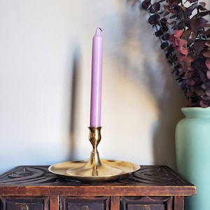 May include: A brass candle holder with a purple candle on a wooden table with drawers. The candle holder has a round base and a tall, slender stem.