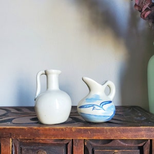 May include: Two small white ceramic pitchers with blue and white painted designs. The pitcher on the left is plain white, while the pitcher on the right has a blue and white bird design.