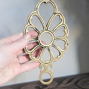 May include: A gold-toned metal trivet with a decorative floral design. The trivet has a circular opening in the center and a loop at the bottom.