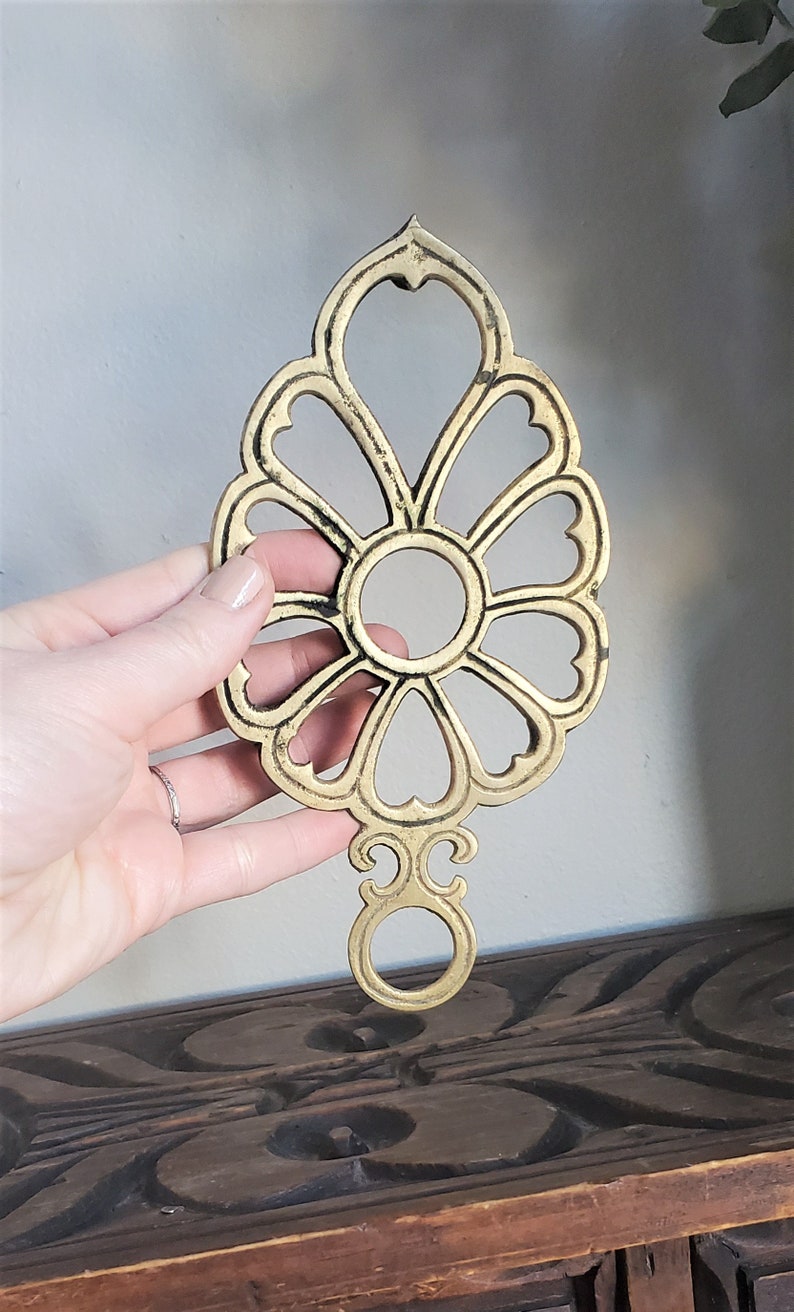 May include: A gold-toned metal trivet with a decorative floral design. The trivet has a circular opening in the center and a loop at the bottom.