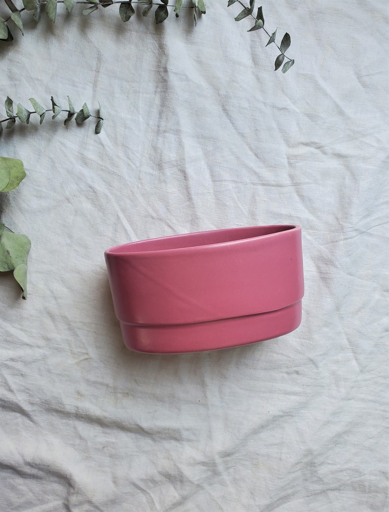 May include: A pink oval ceramic planter with a smooth, glossy finish. The planter is sitting on a white fabric surface.