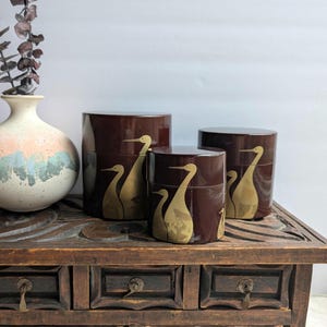 May include: Three dark brown, cylindrical containers with gold crane designs. The containers are of varying heights and sit on a wooden table with drawers. A decorative vase with a pink and blue design is to the left.