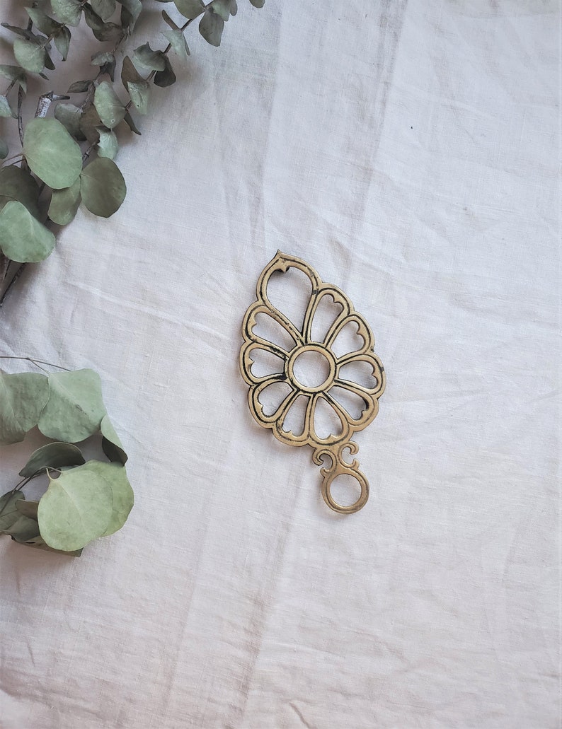 May include: A gold-toned metal decorative object with a floral design. The object has a round center and eight petals that are shaped like hearts. The object has a small loop at the bottom.