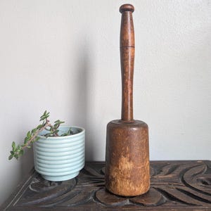 Puede incluir: Un mortero de madera vintage con una base cilíndrica y un mango largo. El mortero es de color marrón cálido y muestra signos de desgaste. Se encuentra sobre una superficie de madera oscura junto a una pequeña maceta de cerámica azul claro con una planta.