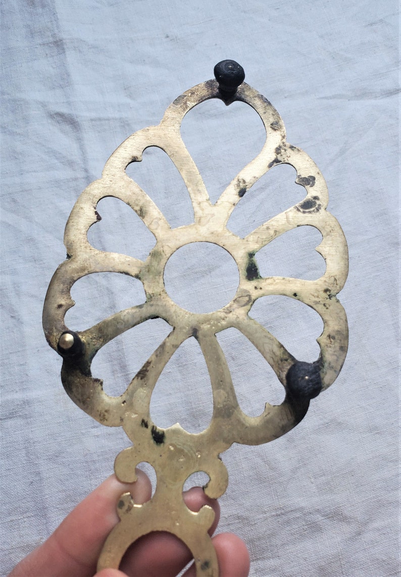May include: A brass trivet with a decorative floral design and three black feet. The trivet is designed to protect surfaces from hot pots and pans.