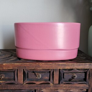 May include: A pink ceramic planter with a smooth, glossy finish. The planter has a rounded rectangular shape with a slightly raised lip around the top edge.